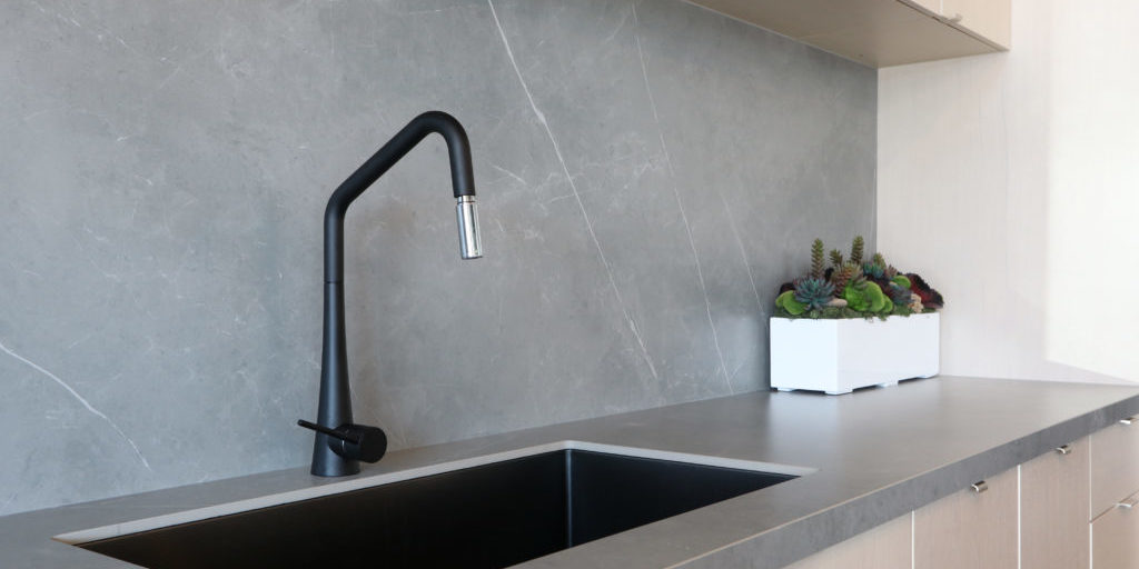 A black sink and matt finish black faucet set against a grey countertop and backsplash made of porcelain slabs that mimic the natural look of stones, decorated by green indoor planter.